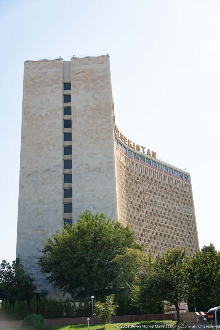 Close upward view of Hotel Uzbekistan's curved screened facade and rooftop UZBEKISTAN letters.