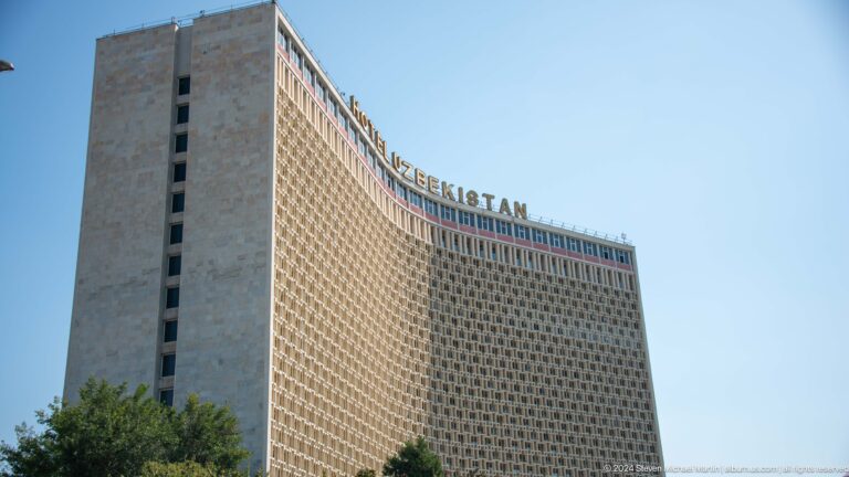 Full curved facade of Hotel Uzbekistan with geometric screening and rooftop lettering under a blue sky.