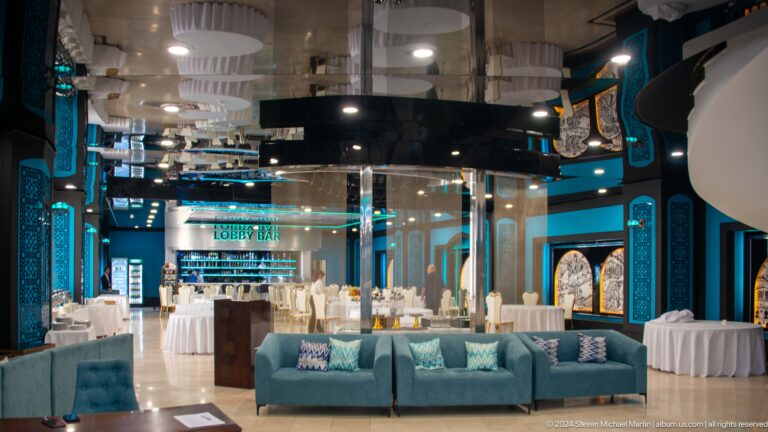 Teal hotel lobby with curved sofas, glass enclosure, banquet tables, and a LOBBY BAR sign.