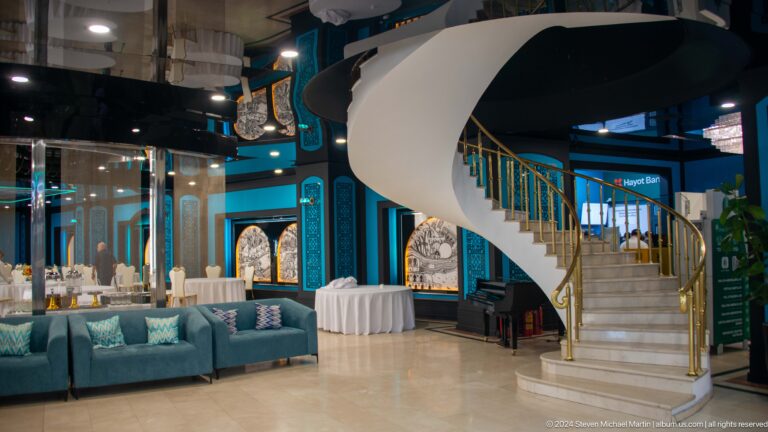 White spiral staircase with gold railings curving through a teal hotel lobby beside a piano.