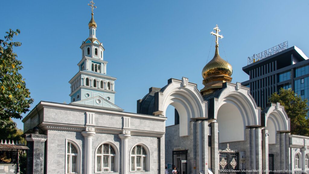 Pale blue cathedral bell tower, arched entrance, and gilded onion domes beneath a clear sky.