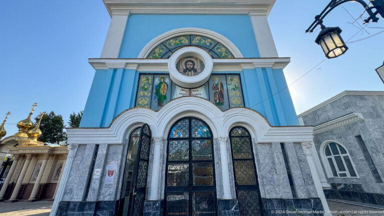 Blue cathedral entrance with arched doors, saint mosaics, and a circular image of Christ.