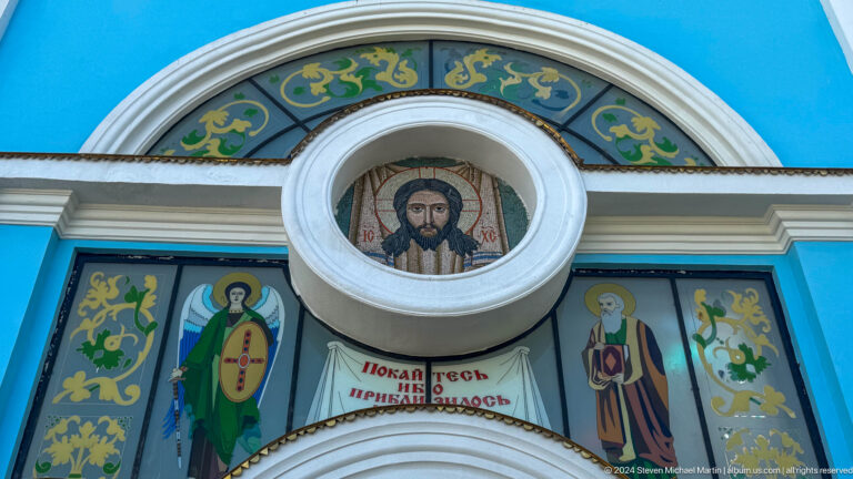 Circular image of Christ above saint mosaics and Cyrillic text on a blue facade.