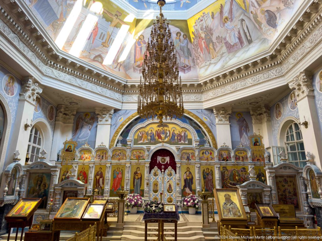 Rich cathedral interior with gilded iconostasis, chandelier, painted dome, icons, and flowers.