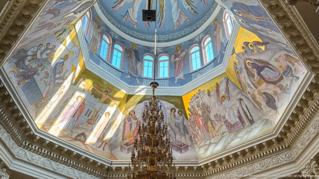Upward view of a blue cathedral dome painted with angels and saints above a chandelier.