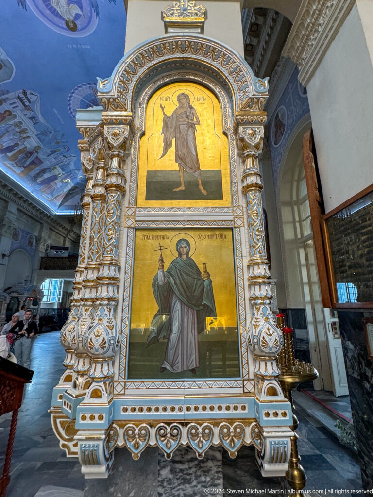 Full-length icon of a female saint holding a cross in an ornate carved frame.