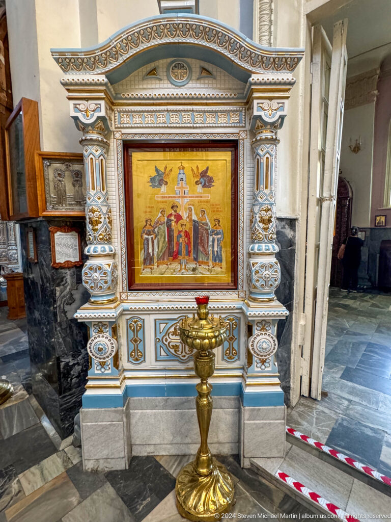 Framed gold icon of saints beneath a cross and angels, with a votive lamp below.
