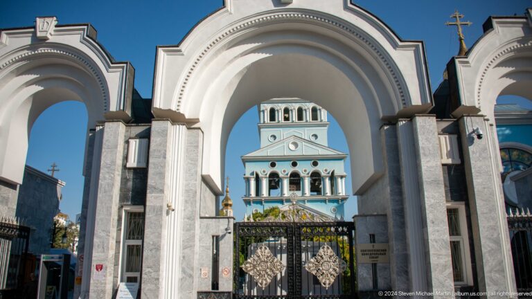 White cathedral gateway framing a black iron gate and pale blue bell tower.