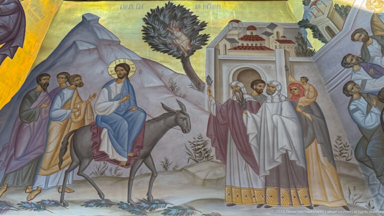 Mural of Christ riding a donkey toward a city gate as crowds gather.