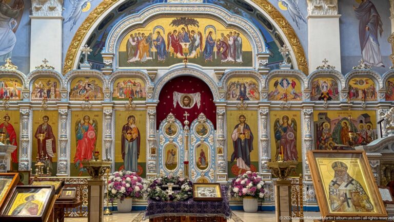 Frontal gilded iconostasis with royal doors, saint icons, Last Supper panel, and flowers.