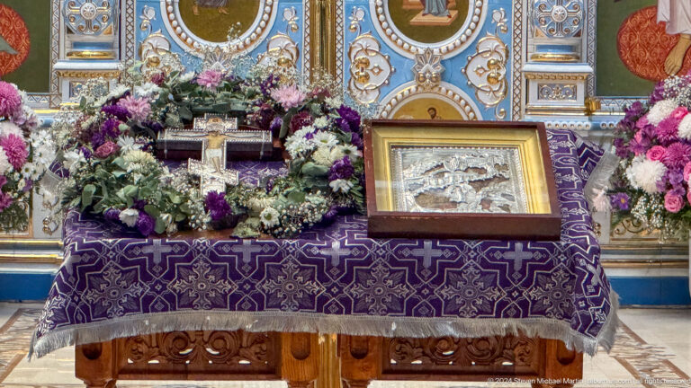 Purple-covered devotional table with flowers, cross, framed sacred objects, and book.