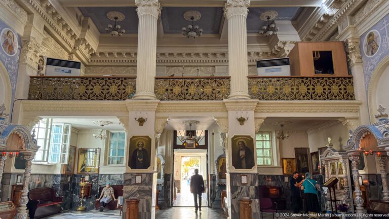 Tall cathedral hall with white columns, gold balcony rail, wall icons, and open doors.