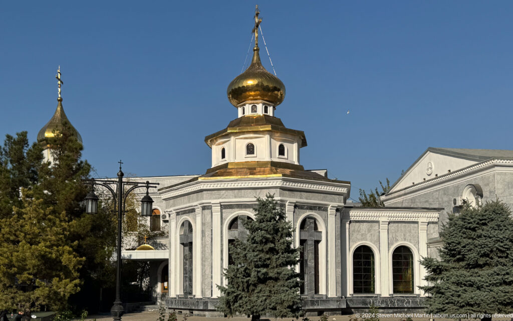 Octagonal pale church building with arched windows and a gilded onion dome among trees.