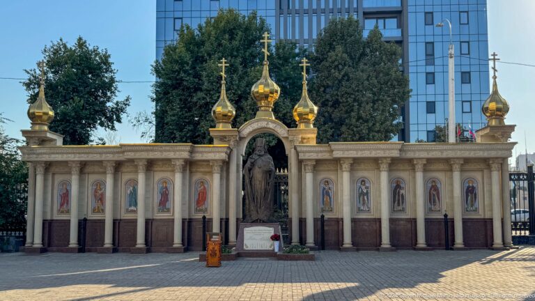 Long icon-lined cathedral wall with a central niche and three gilded domes beneath modern buildings.