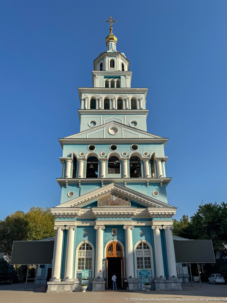 Frontal pale blue bell tower with stacked arcades, clock, and triangular mosaic.