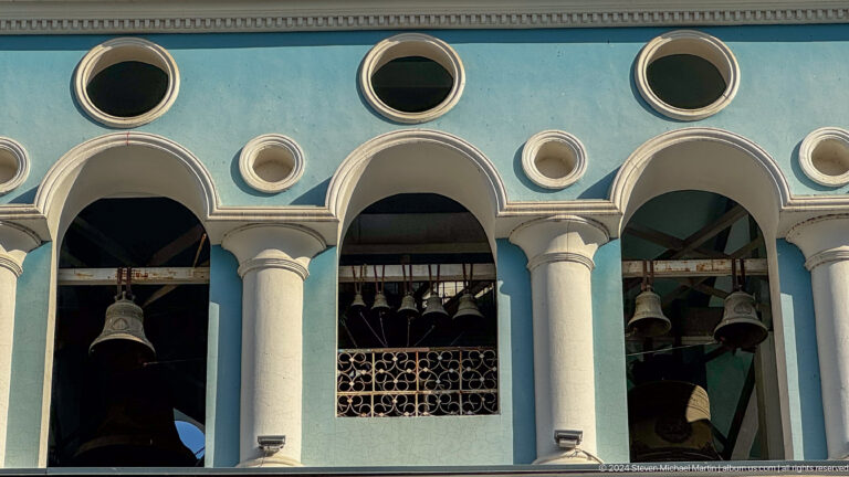 Close view of bells behind white columns and arches in a pale blue bell tower.