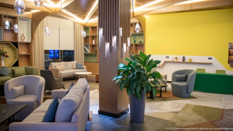 Modern hotel lounge with gray seating, wood paneling, a yellow wall, built-in shelves, and a tall potted plant.