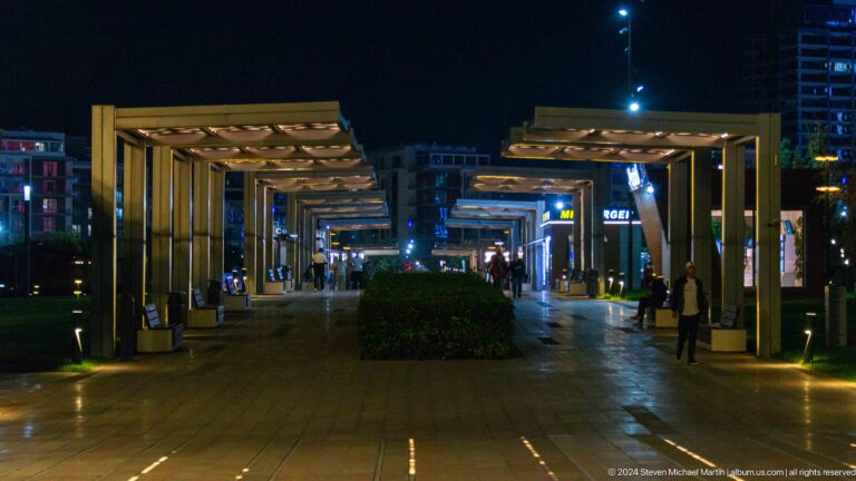Night plaza with a circular sculpture, international flags, fountains, pedestrians, and brightly lit towers.