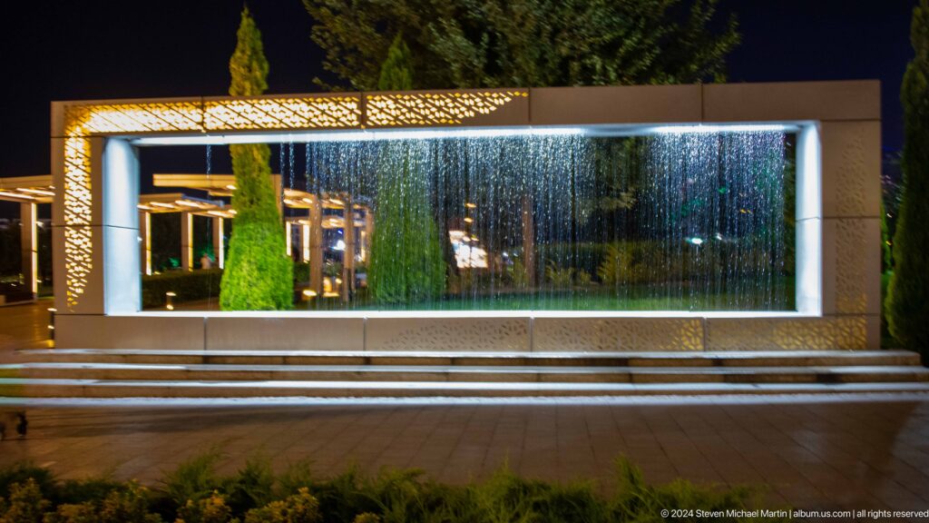 Low fountain spray in a dark park foreground with blue-, red-, and green-lit towers in the distance.