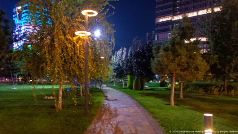 Nighttime park path beside a bright linear feature, with trees, shrubs, and distant high-rise towers.