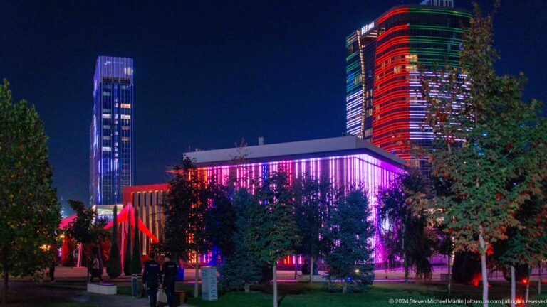 Dark landscaped foreground with a long bright strip and blue-, red-, and green-lit towers beyond.