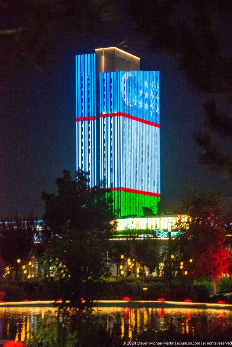 Night view of a pale multi-story building with repeated windows, balconies, warm interior lights, trees, and parked vehicles.