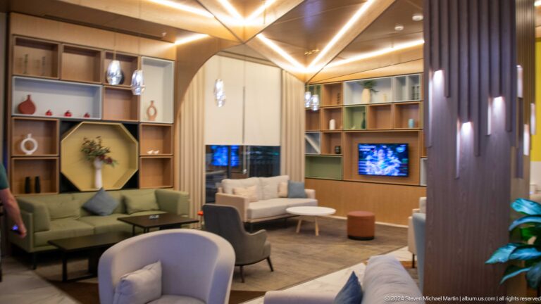 Hotel lounge with a curved wood partition, geometric ceiling lights, built-in shelves, sofas, chairs, and a television.