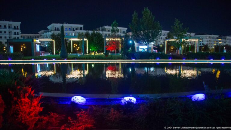 Reflecting pool at night with illuminated pergolas, trees, and low-rise buildings mirrored in the water.