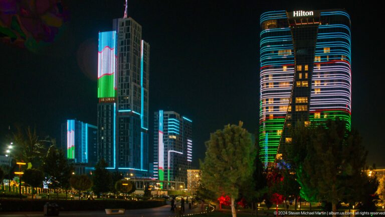 Wide night skyline with Hilton signage and several towers illuminated in Uzbekistan's national colors.