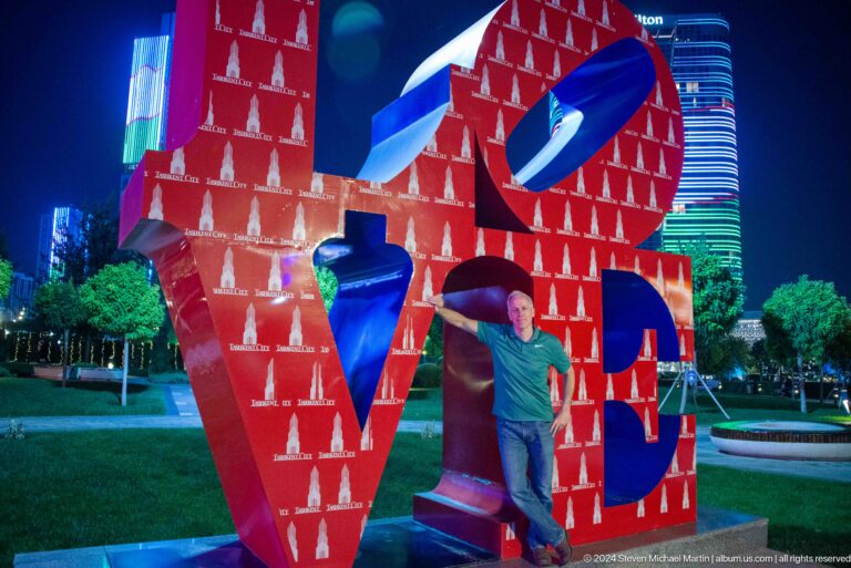Man posing beside a large red LOVE sculpture patterned with white tower motifs at night.