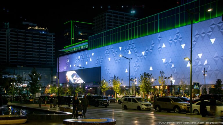 Busy night street outside Tashkent City Mall with a white triangular-panel facade and green roof lighting.