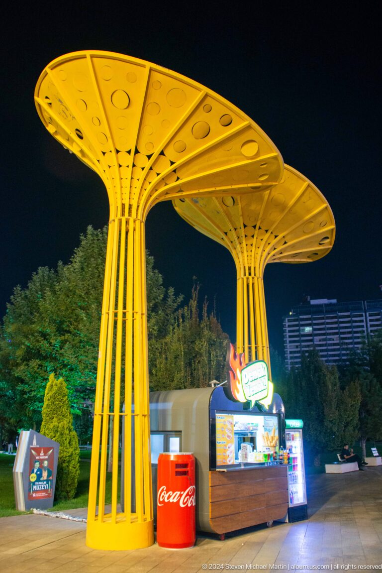 Yellow tree-like metal canopies with branching supports and wide perforated roofs at night.