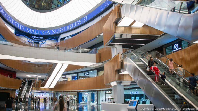 Multi-level shopping mall atrium with escalators, curved wood-clad balconies, retail stores, and a bright ceiling.