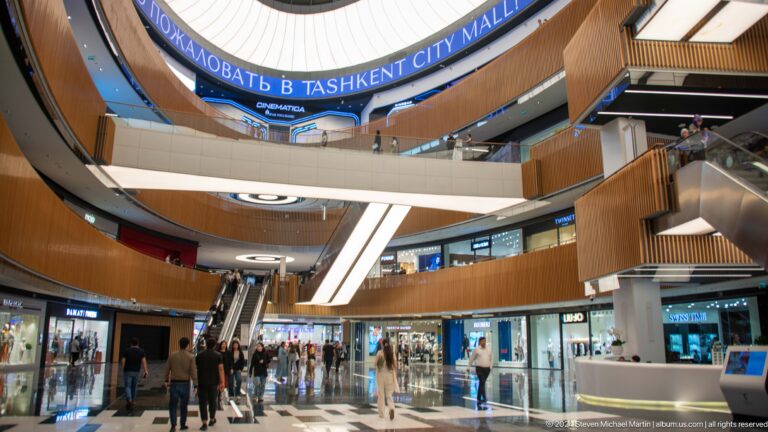 Shoppers crossing a bright mall atrium beneath escalators and bridges with wood-clad balconies.