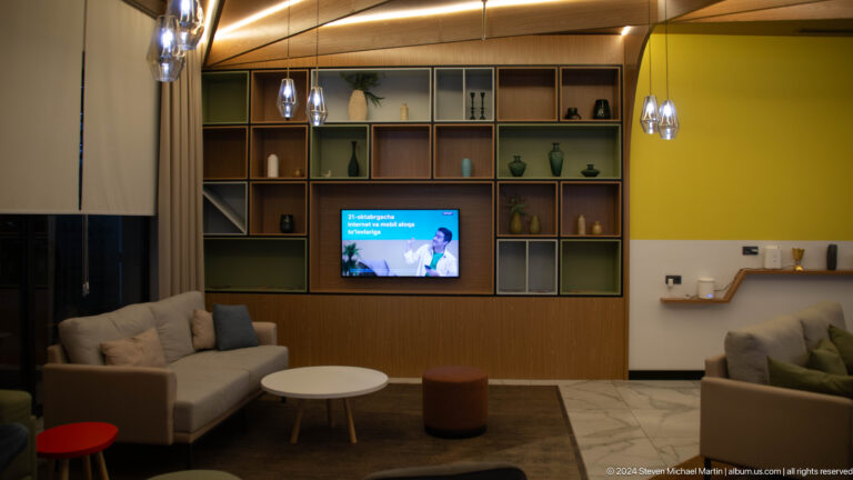 Grid-style built-in shelving with decorative vessels and a television above a sofa and round table in a hotel lounge.