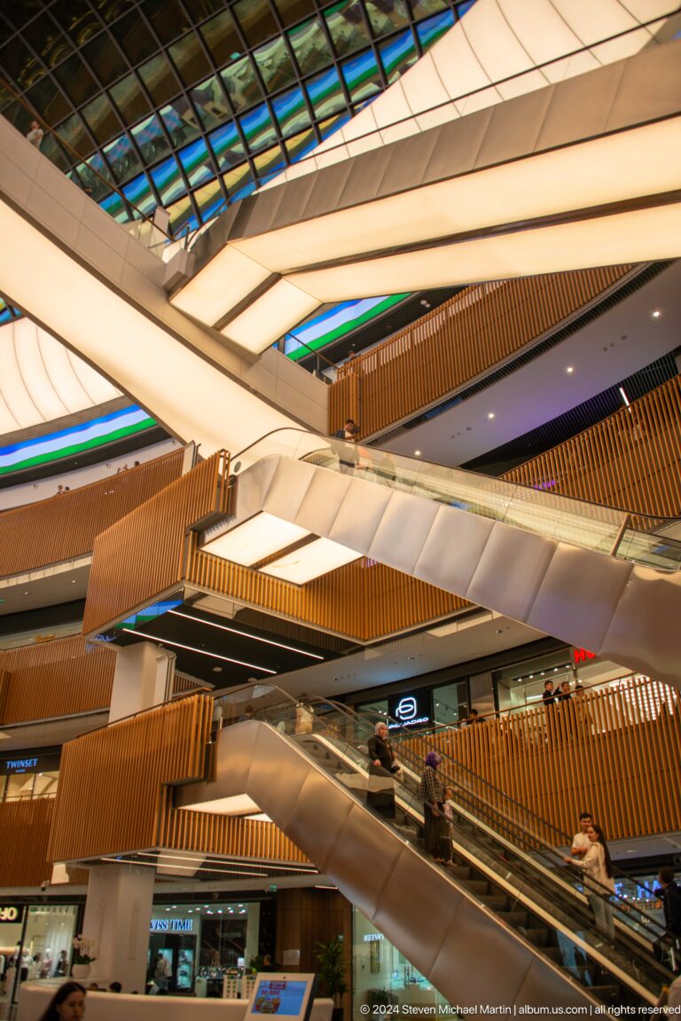 Upward view of crossing escalators, wood-clad balconies, glass railings, and bright ceiling panels.