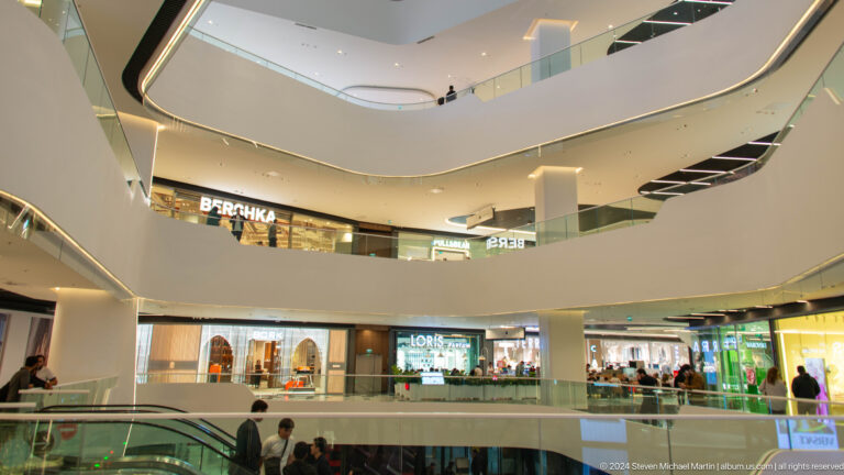 Bright mall interior with curved white balconies, glass rails, fashion stores, and visitors below.