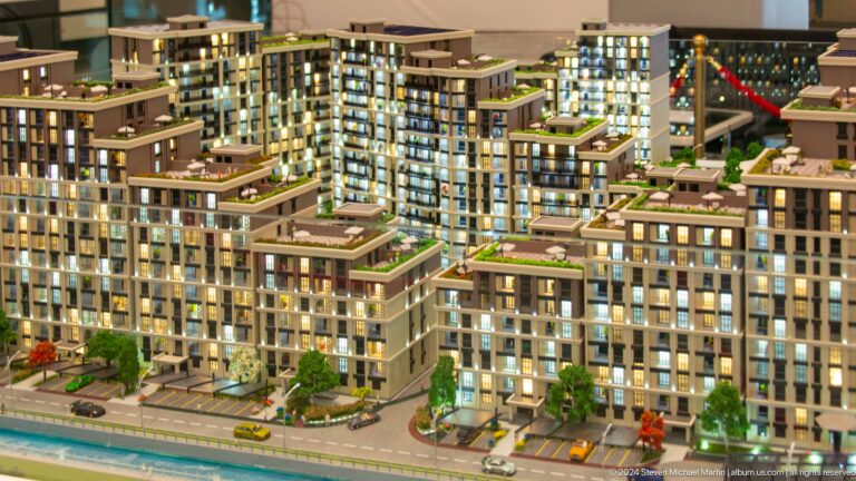 Illuminated architectural model of terraced apartment buildings with green roofs, streets, trees, and miniature cars.