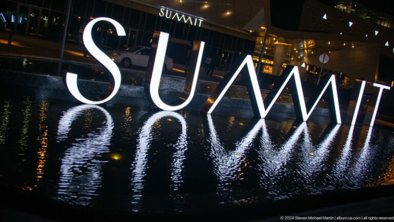 Large glowing SUMMIT letters reflected in a shallow pool outside a dark building at night.