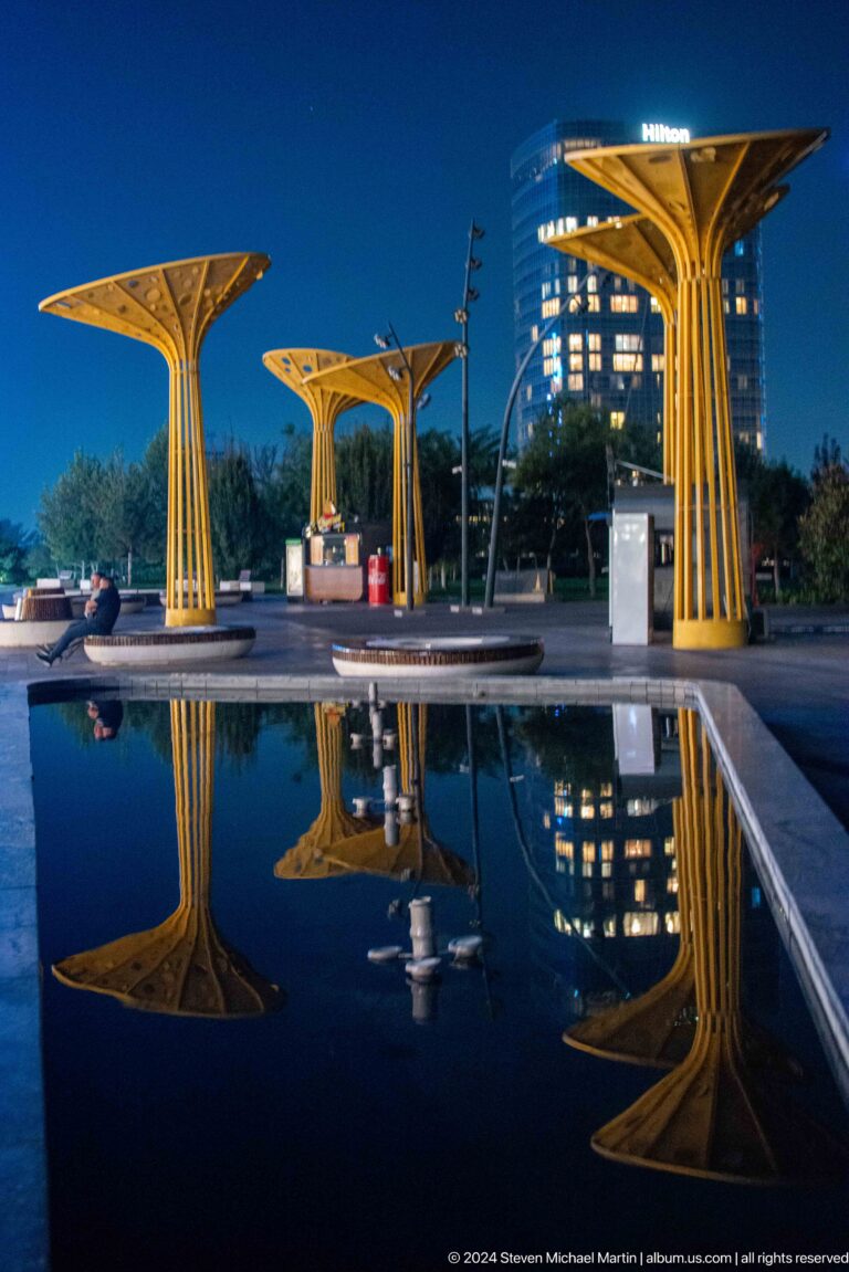 Yellow tree-like canopies and round benches reflected in a still park pool at dusk.