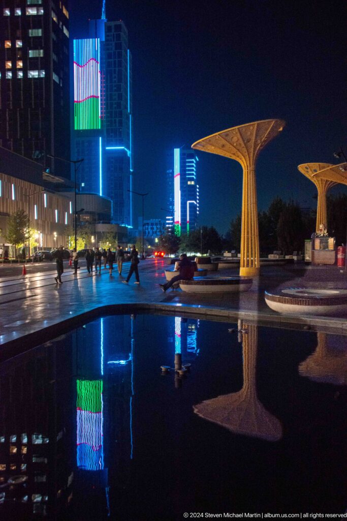 Pedestrians crossing wet pavement beside yellow canopies and a reflecting pool with blue-lit towers beyond.