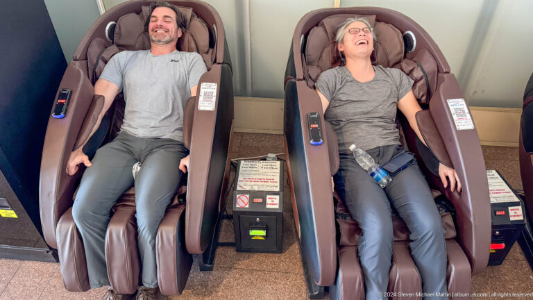 Two adults reclining and laughing in large brown massage chairs inside a mall.