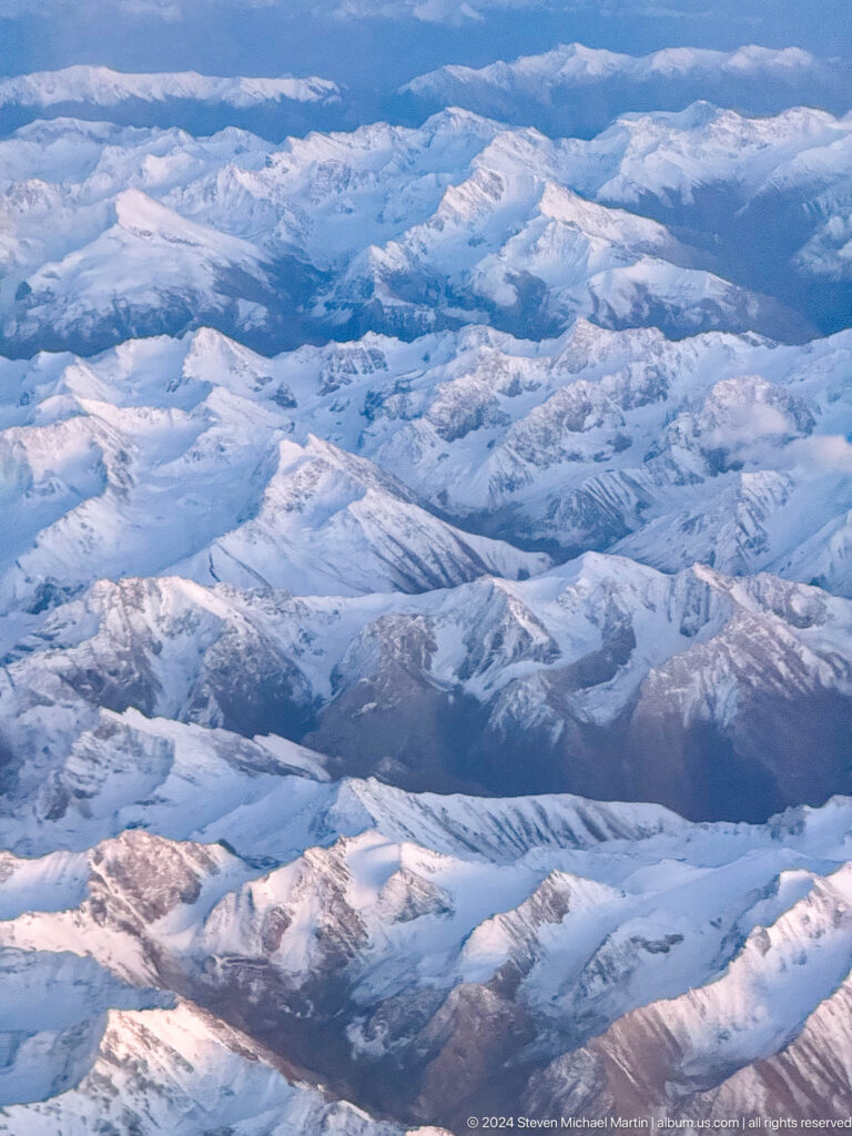 Aerial view of rugged snow-covered mountains, ridges, and deep shadowed valleys.