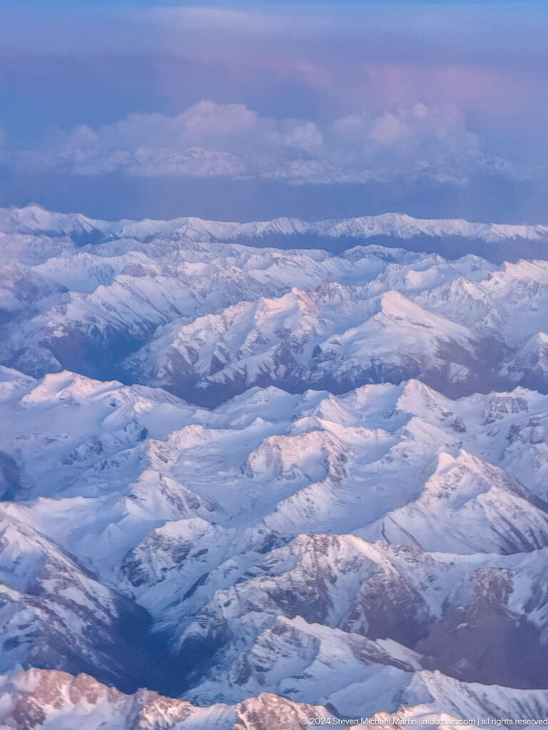 Aerial view of layered snow-covered mountain chains and valleys fading into blue haze.