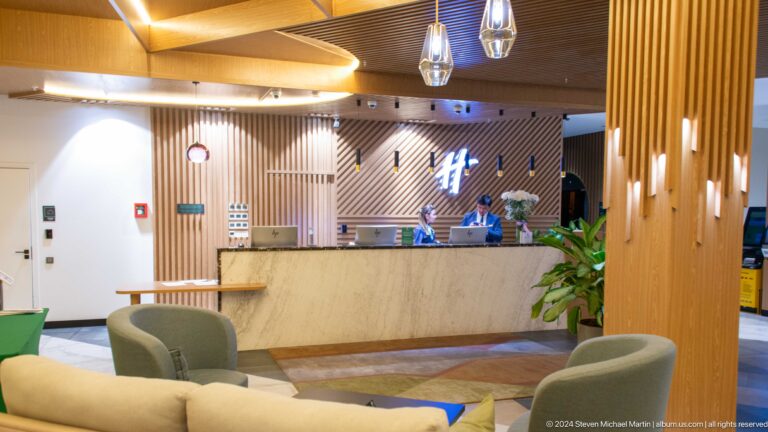Hotel reception desk with two staff members, pendant lights, a pale counter, and an illuminated logo on a slatted wood wall.