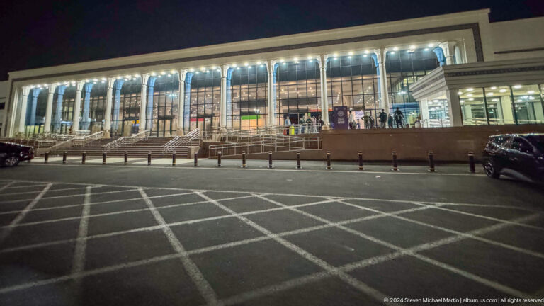 Night view of a large glazed airport terminal with stairs, ramps, passengers, and a striped drop-off roadway.