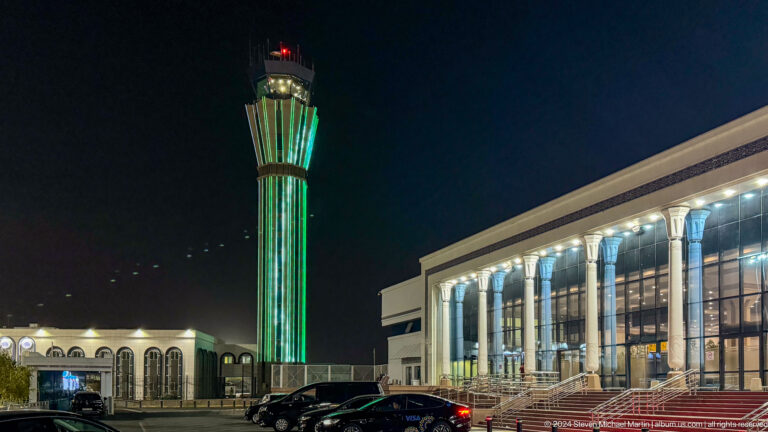 Green-lit airport control tower beside a bright terminal building and curbside cars at night.