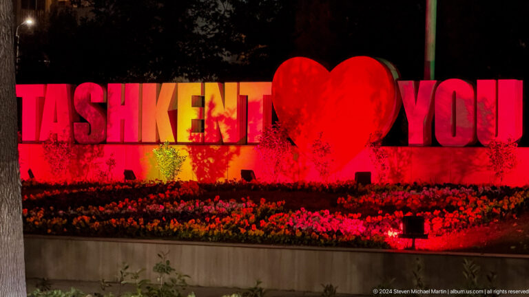 Large red Tashkent I Love You sign with a heart symbol behind a flower bed at night.