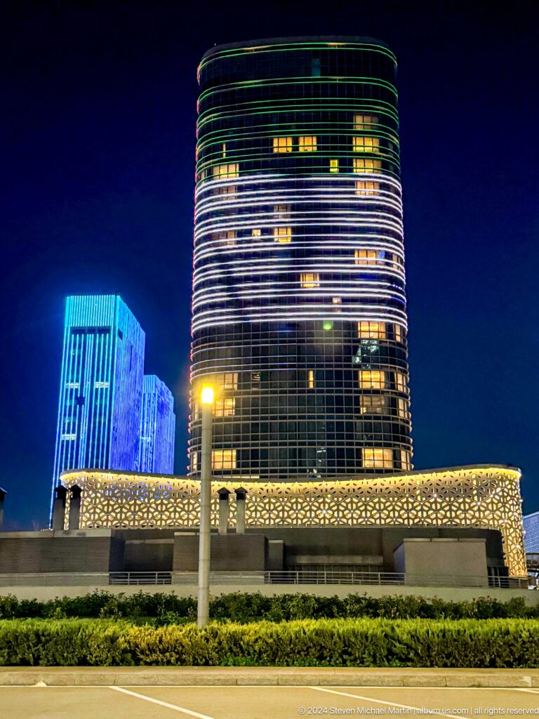 Curved Hilton tower above a gold geometric facade with a blue-lit high-rise behind at night.