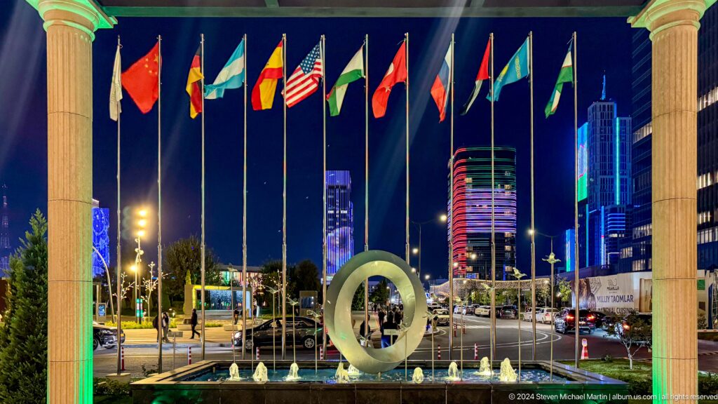 International flags above a circular sculpture and small fountains with brightly lit towers behind at night.